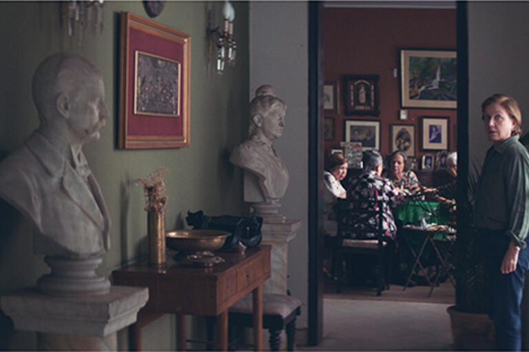 Woman standing at the end of a hallway, behind her there is a round table with four woman playing cards