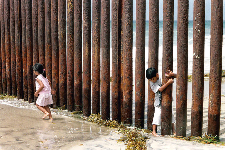 Children play through the U.S.–Mexico border wall made of vertical steel beams on the beach near San Diego in 2001. (Photo by Quim Gil.)