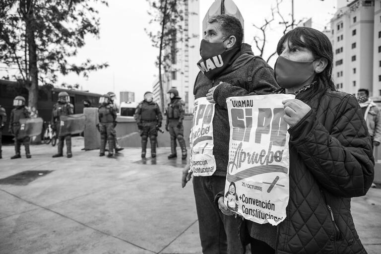 Standing in front of police in riot gear, a masked mature couple with signs supports the campaign for  a new constitution in Chile,  August 2020. (Photo by Paulo Slachevsky.)