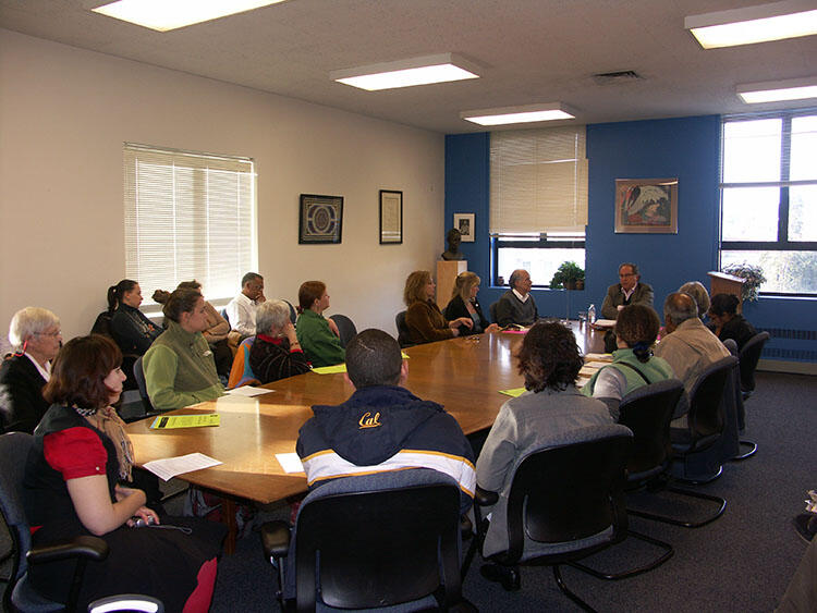 The speaker is speaking to a group of people gathered around a large table in a meeting room.