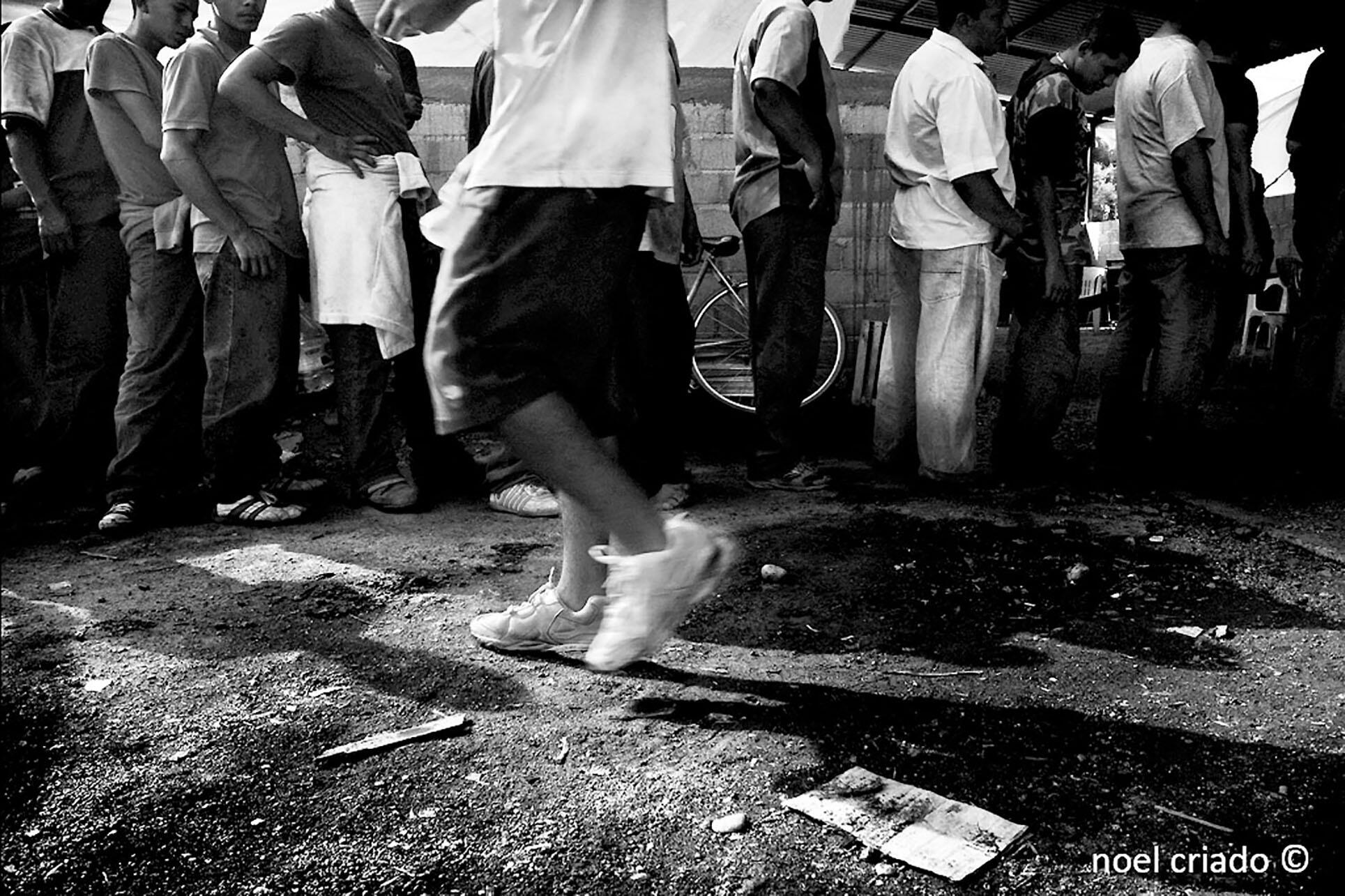 A black and white photo of a line of men waiting at Hermanos en el Camino, a migrant shelter in Ixtepec, Oaxaca. (Photo by Noel Criado.)