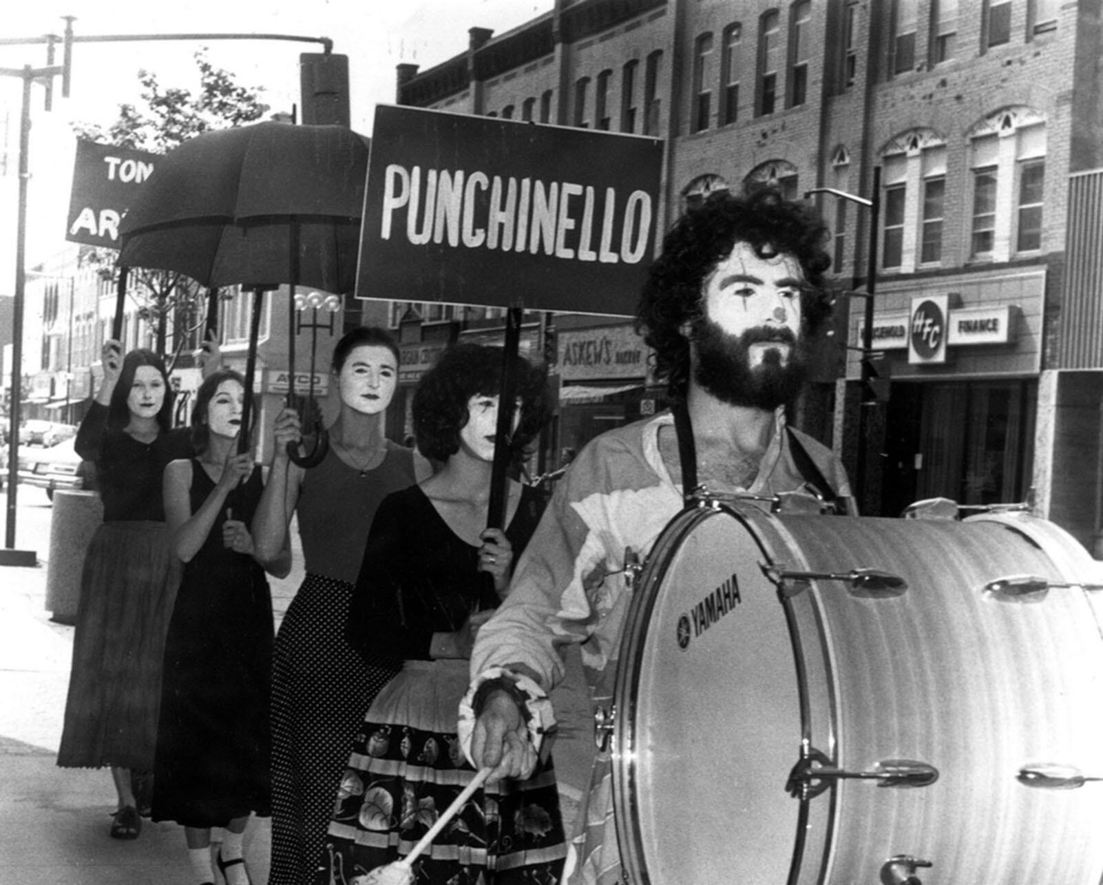 A traveling theater group walks in makeup behind a drum down a street in a small town. (Photo courtesy of Elgin County Archives.)